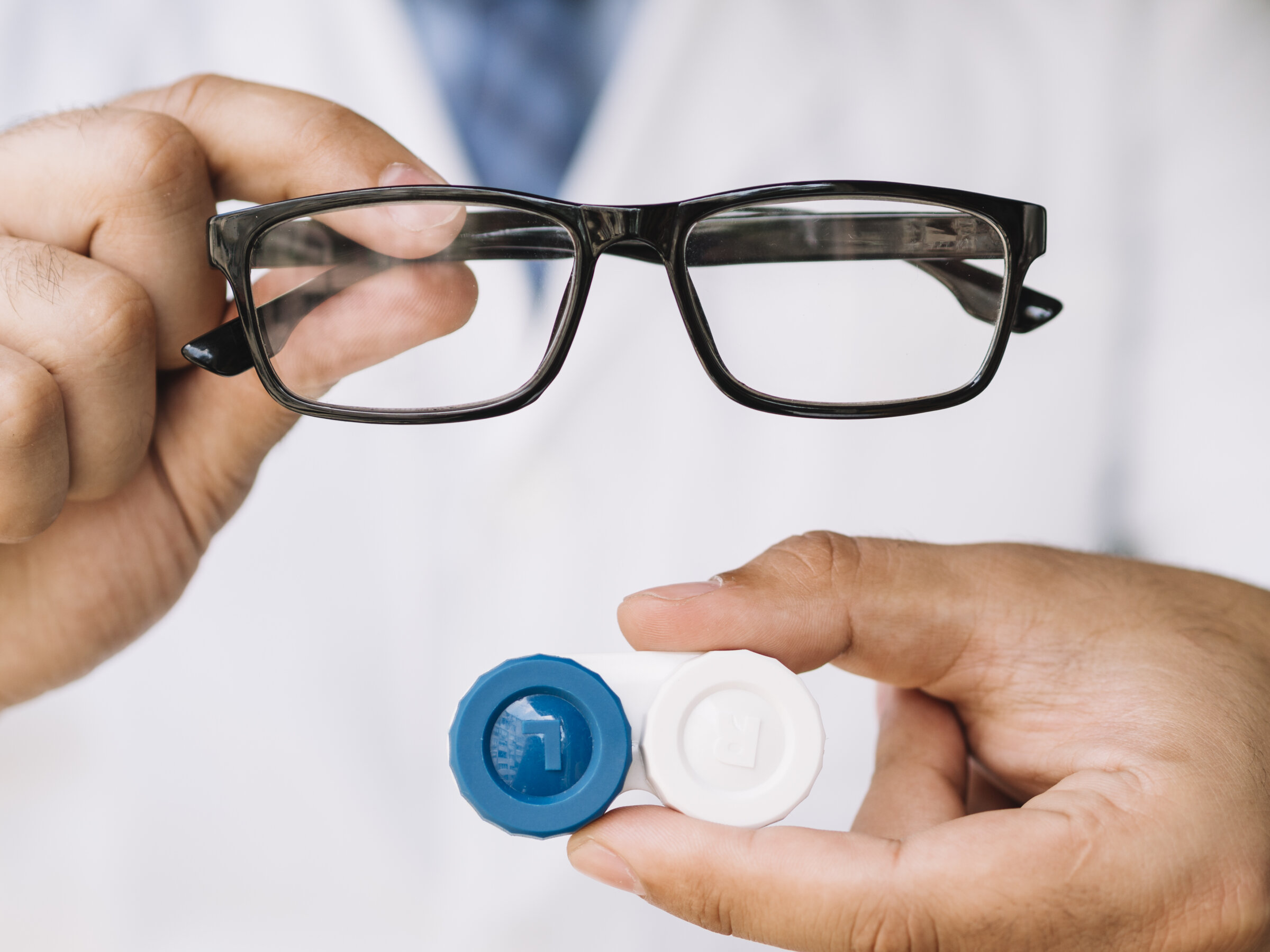 Brille oder Kontaktlinsen? Entscheidungshilfe von Optik Stöckert Nahaufnahme: Person hält eine schwarze Brille in der einen Hand und ein Kontaktlinsenbehälterpaar in der anderen – Entscheidungshilfe zwischen Brille und Kontaktlinsen.