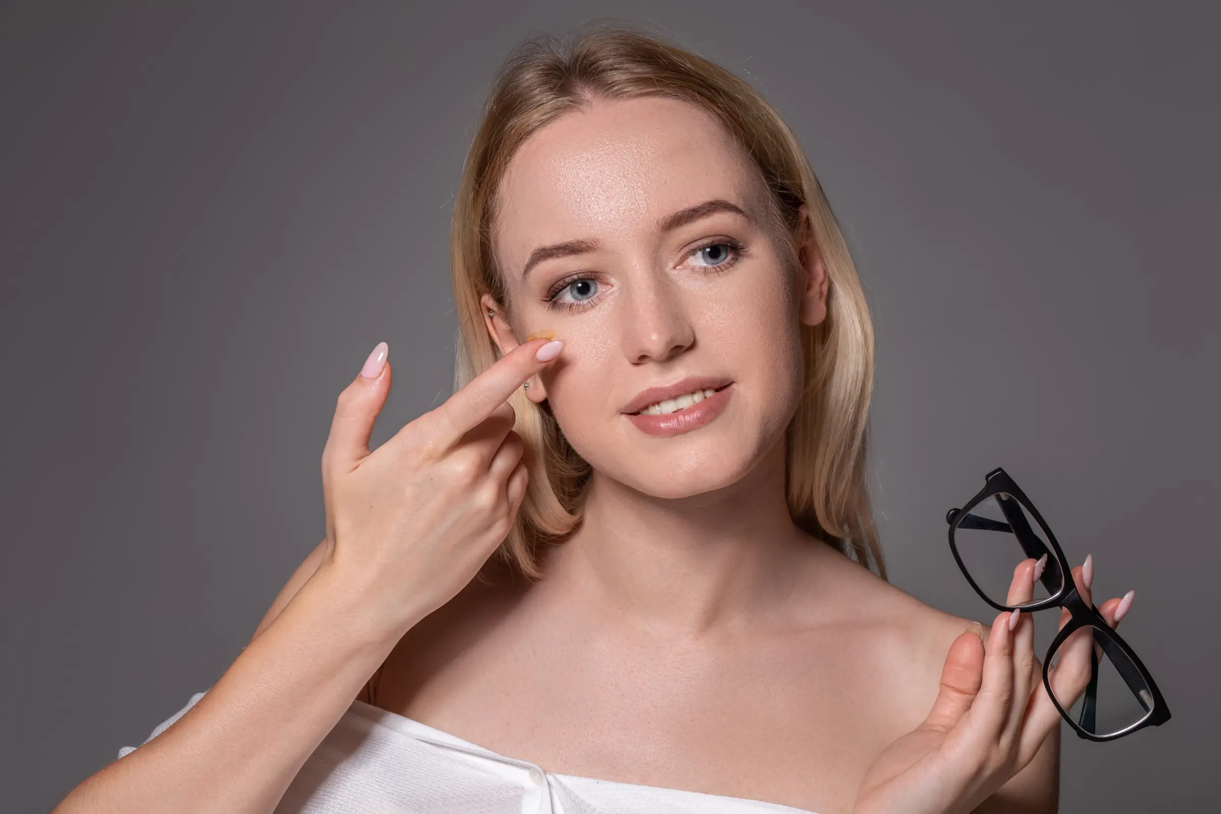 Junge Frau mit blonden Haaren hält eine Kontaktlinse nah ans Auge und eine Brille in der anderen Hand – sinnbildlich für die Entscheidung zwischen klassischer Brille und innovativer Nachtlinse wie DreamLens.