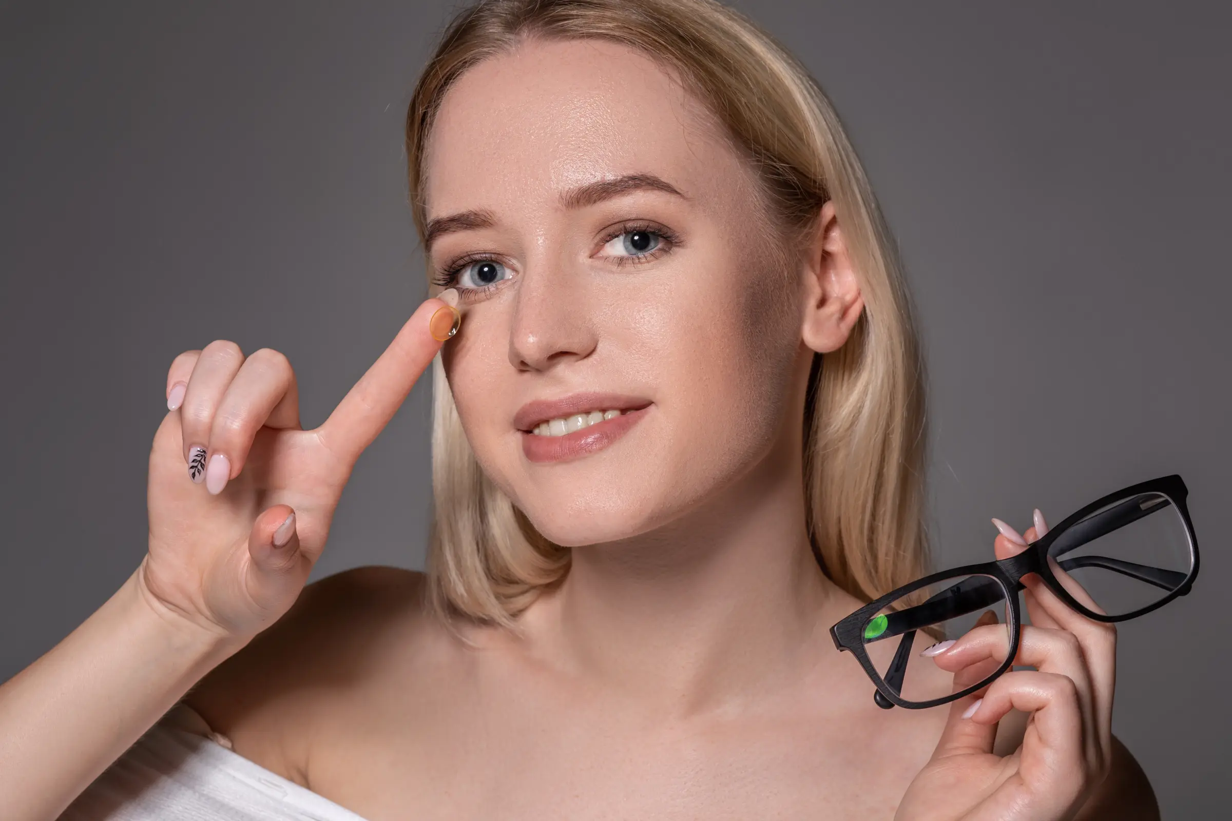 Junge Frau mit blonden Haaren hält eine Kontaktlinse am Finger und eine Brille in der anderen Hand – Symbolbild für die Entscheidung zwischen Brille und Dreamlens-Nachtlinsen.